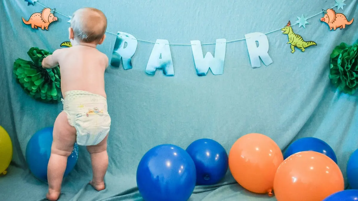 baby in white diaper standing beside blue and red balloons